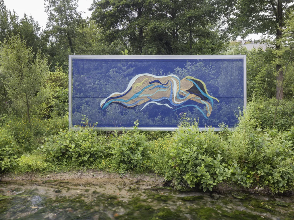 Ein großformatiges Plakat aus Stoff mit einer abstrakten, wellenförmigen Figur in Blau-, Beige- und Grüntönen steht inmitten dichter Vegetation am Wasser. Die Figur scheint über das leuchtend blaue Hintergrundmotiv zu schweben, das den Himmel und Bäume reflektiert.