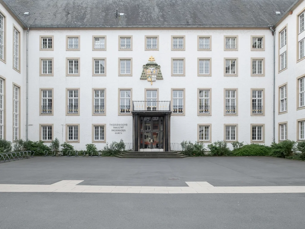 Das Bild zeigt die Vorderseite eines historischen Gebäudes mit symmetrisch angeordneten Fenstern und einem zentralen Eingang. Über dem Eingang befindet sich ein Wappen und die Inschrift "Theologische Fakultät Paderborn."