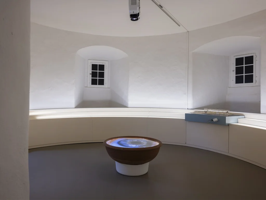 Ein heller, runder Raum mit weiß verputzten Wänden zeigt in der Mitte eine Projektion eines Augapfels in einer schalenartigen Installation. Umgeben ist der Raum von einer weißen Sitzbank und einem kleinen Architekturmodell auf einem Podest.