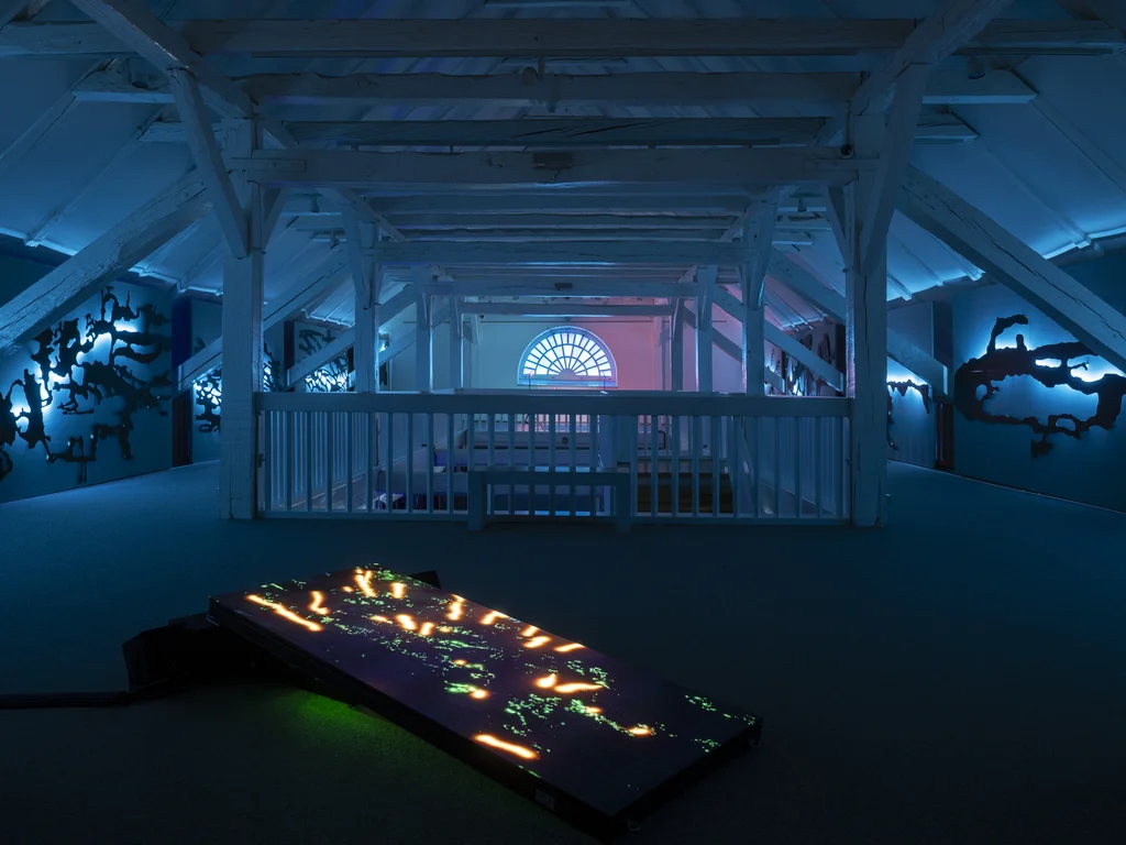 Das Bild zeigt einen Raum mit einer Holzdecke, beleuchtet durch blaue Lichter, mit einem beleuchteten rechteckigen Objekt im Vordergrund und Schattenfiguren an den Wänden.